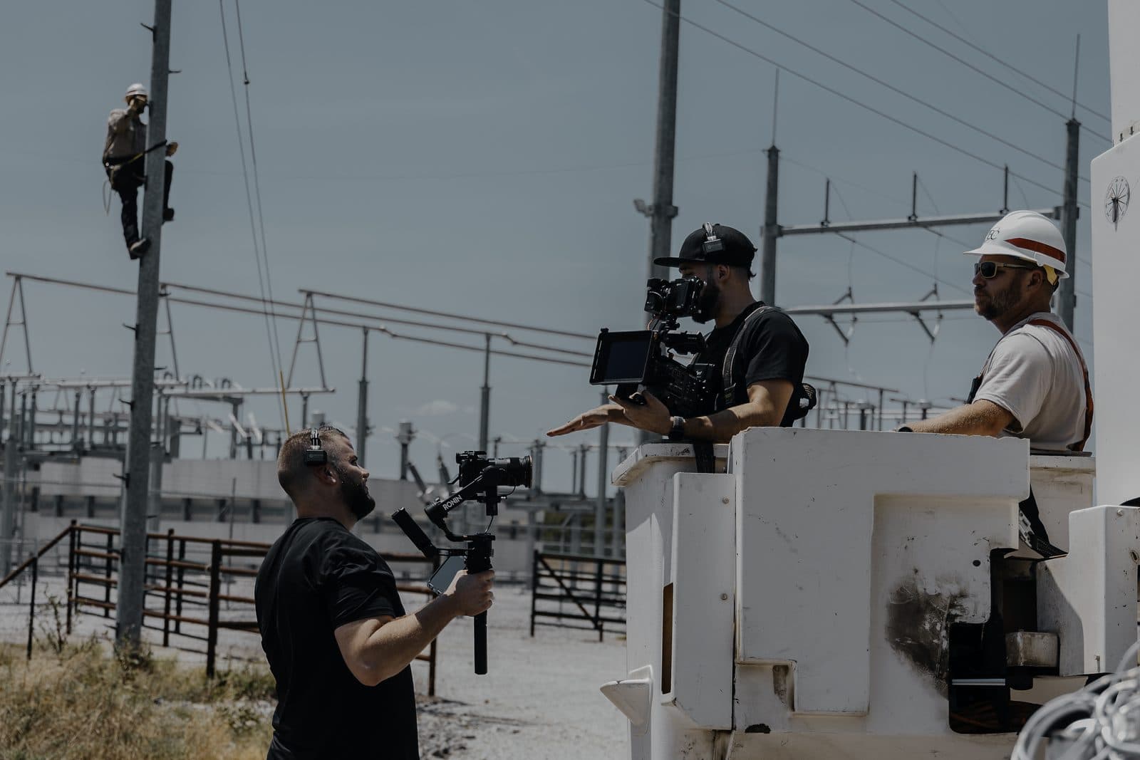A Hyped Visuals camera operator films People's Electric Cooperative crew members from a bucket truck at a substation, with a lineman climbing a utility pole in the background, Ada, Oklahoma